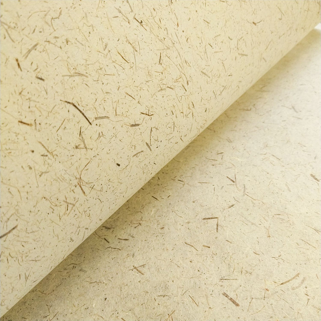 A close-up of Pine Leaf Kozo Paper by Kozo Studio showcases its beige color with natural fibers and texture. The paper rolls diagonally across the image, highlighting its organic look and subtle details of traditional rice paper.