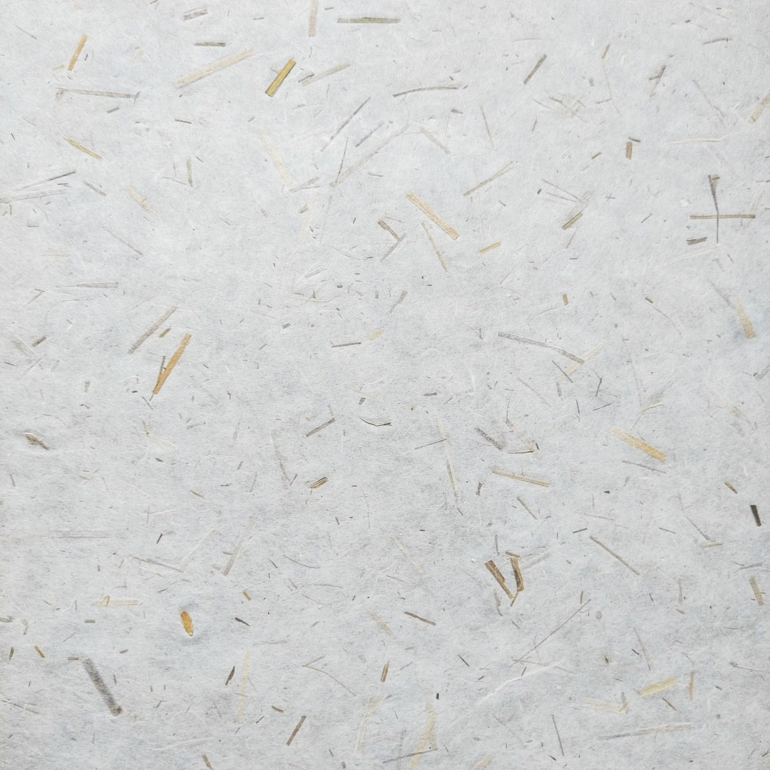 A close-up of Rice Straw Confetti Kozo Paper by Kozo Studio shows textured rice paper with straw-like fibers scattered across a white surface, creating an organic, rustic look with varying beige shades.