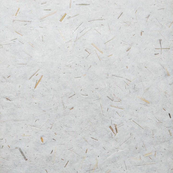 A close-up of Rice Straw Confetti Kozo Paper by Kozo Studio shows textured rice paper with straw-like fibers scattered across a white surface, creating an organic, rustic look with varying beige shades.