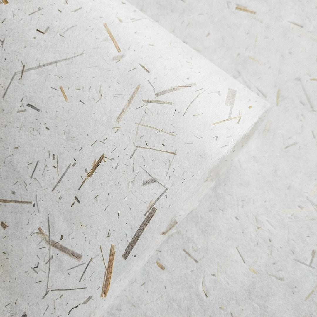A close-up showcases Rice Straw Confetti Kozo Paper by Kozo Studio, featuring textured mulberry paper with embedded straw fibers. These fibers vary in length and color, creating an organic pattern against the white background. One edge of this exquisite paper is slightly curled.