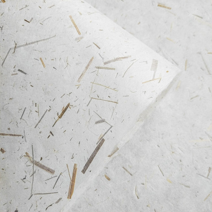 A close-up showcases Rice Straw Confetti Kozo Paper by Kozo Studio, featuring textured mulberry paper with embedded straw fibers. These fibers vary in length and color, creating an organic pattern against the white background. One edge of this exquisite paper is slightly curled.