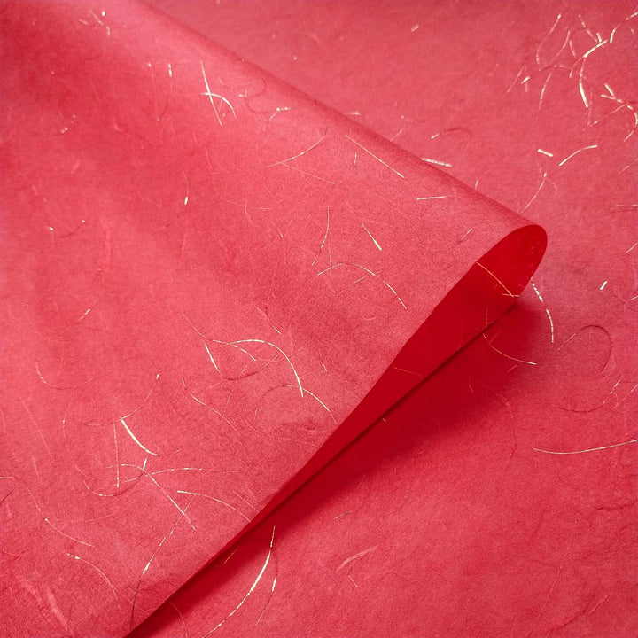 The Golden Tinsel Unryu Kozo Paper (Cherry Red) from Kozo Studio features a slightly textured mulberry surface with delicate gold threads for a shimmering effect, and its corner is gently curled upwards.