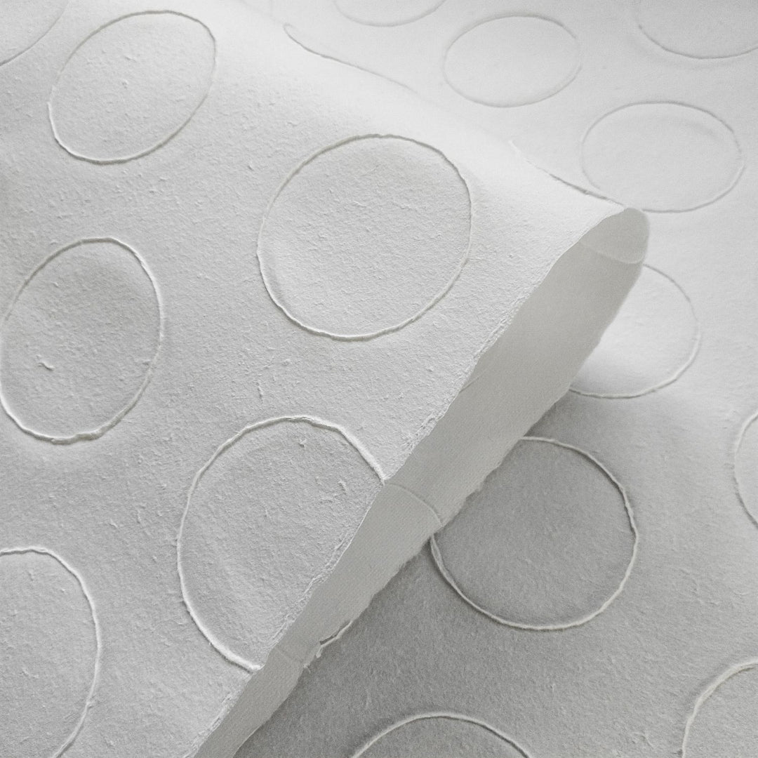 Close-up of Handmade Circle Marking Kozo Paper (White) by Kozo Studio, featuring a textured pattern of raised circles on white embossed mulberry paper. The paper is slightly curled, highlighting its design.