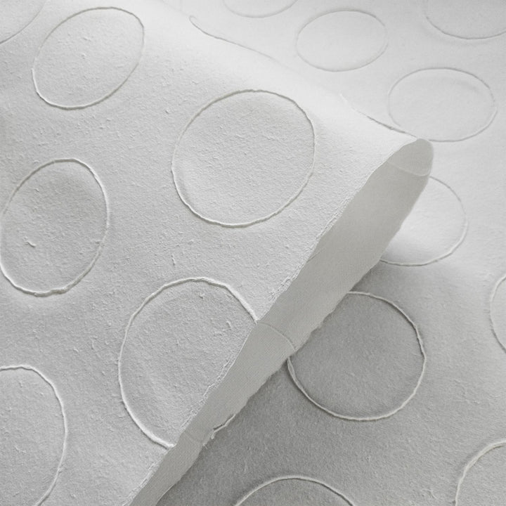 Close-up of Handmade Circle Marking Kozo Paper (White) by Kozo Studio, featuring a textured pattern of raised circles on white embossed mulberry paper. The paper is slightly curled, highlighting its design.