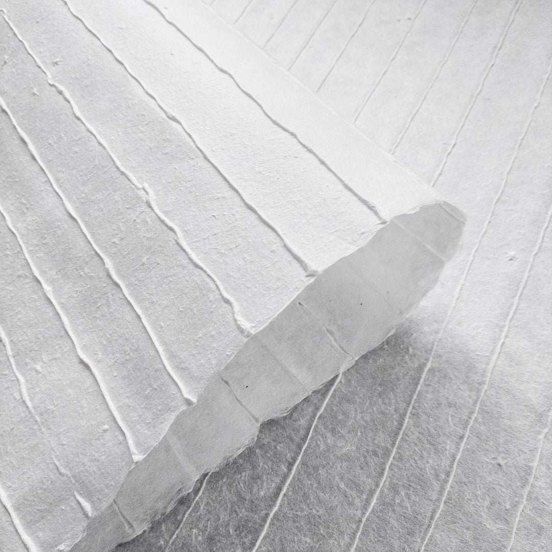 A close-up of Kozo Studios Handmade Stripe Marking Kozo Paper (White) reveals the textured surface with raised vertical lines and its thickness. Partially rolled, it blends seamlessly with the similar background for a uniform appearance.