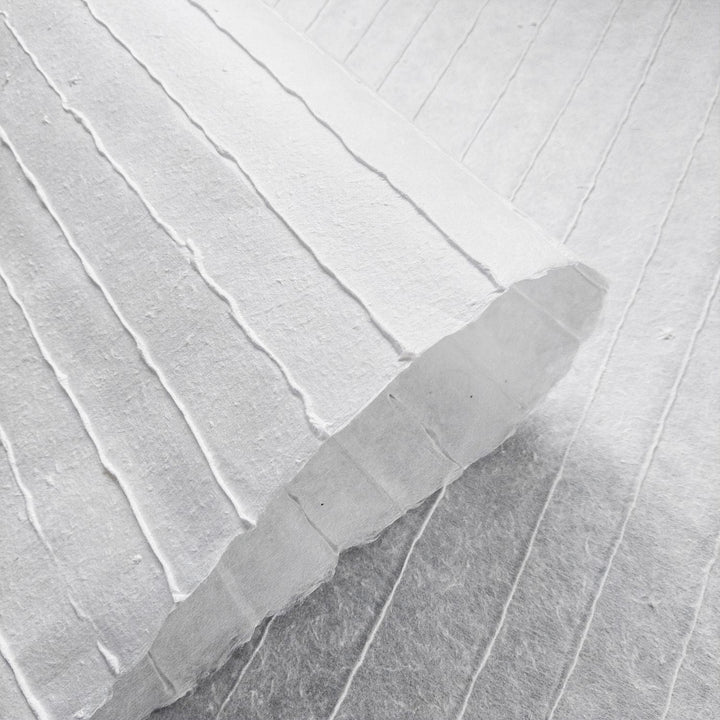 A close-up of Kozo Studios Handmade Stripe Marking Kozo Paper (White) reveals the textured surface with raised vertical lines and its thickness. Partially rolled, it blends seamlessly with the similar background for a uniform appearance.