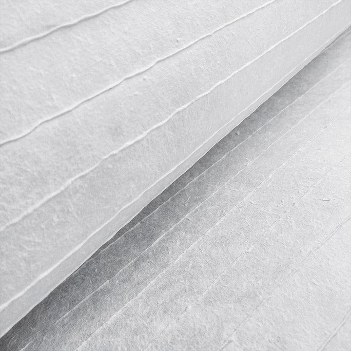 A close-up of Kozo Studios Handmade Stripe Marking Kozo Paper (White) reveals a textured surface with horizontal lines, similar to thick ridged mulberry paper. The lighting highlights the texture and shadows between the lines.
