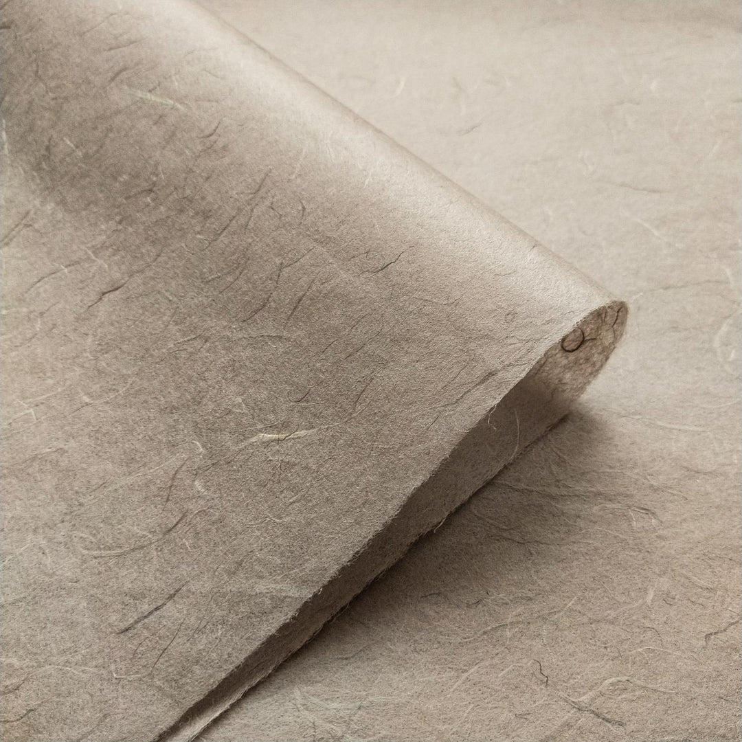 A close-up of a sheet of Kozo Studios Unryu Kozo Paper (Grey), textured and slightly curled at one corner, showcasing its fibrous appearance with visible uneven fibers and a rough surface.