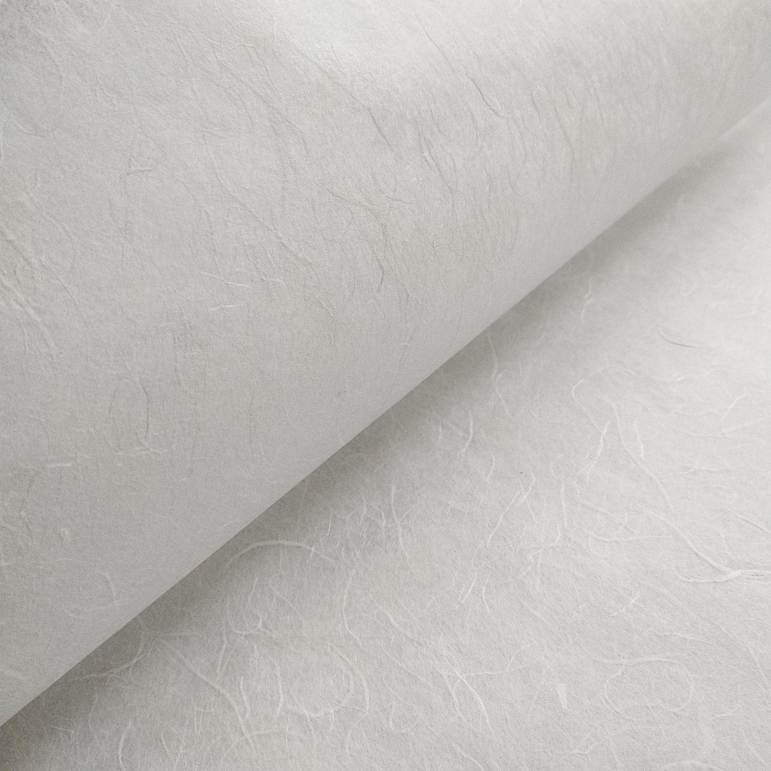 A close-up of Unryu Kozo Paper (White) from Kozo Studio reveals a textured, light gray surface with intricate patterns of embedded fibers, offering a natural, handmade appearance. Slightly rolled, it highlights the papers texture and thickness.