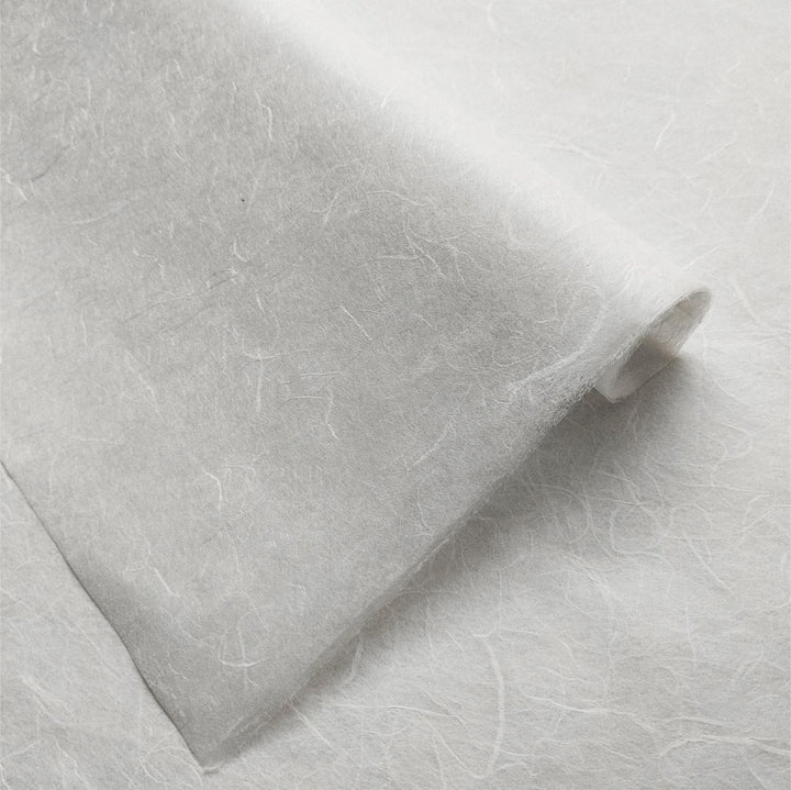 A close-up of Unryu Kozo Paper by Kozo Studio reveals a rich texture with visible fibers. Similar to delicate rice paper, it is partially rolled at one end, showcasing its thin and fragile nature.