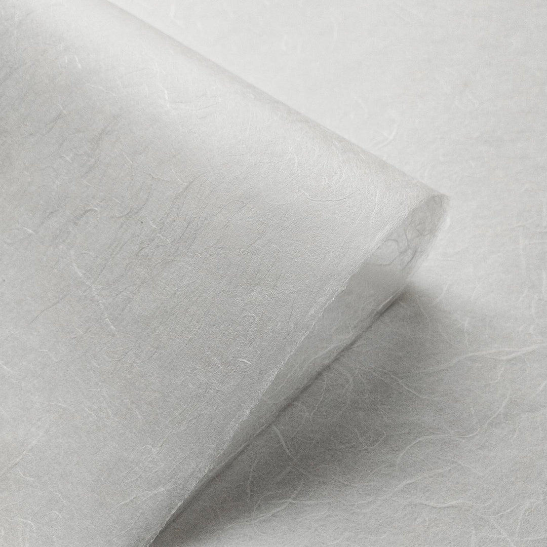 A close-up of Unryu Kozo Paper (White) by Kozo Studio, showcasing its delicate, semi-transparent texture reminiscent of rice paper with textured fibers. The material appears thin and light, with a gentle, soft appearance and slightly curled edges against a smooth surface.