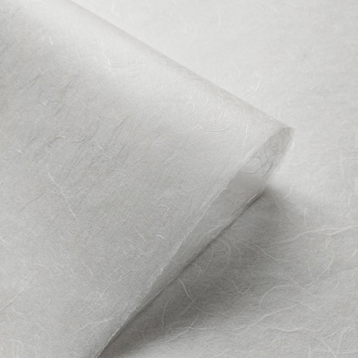 A close-up of Unryu Kozo Paper (White) by Kozo Studio, showcasing its delicate, semi-transparent texture reminiscent of rice paper with textured fibers. The material appears thin and light, with a gentle, soft appearance and slightly curled edges against a smooth surface.