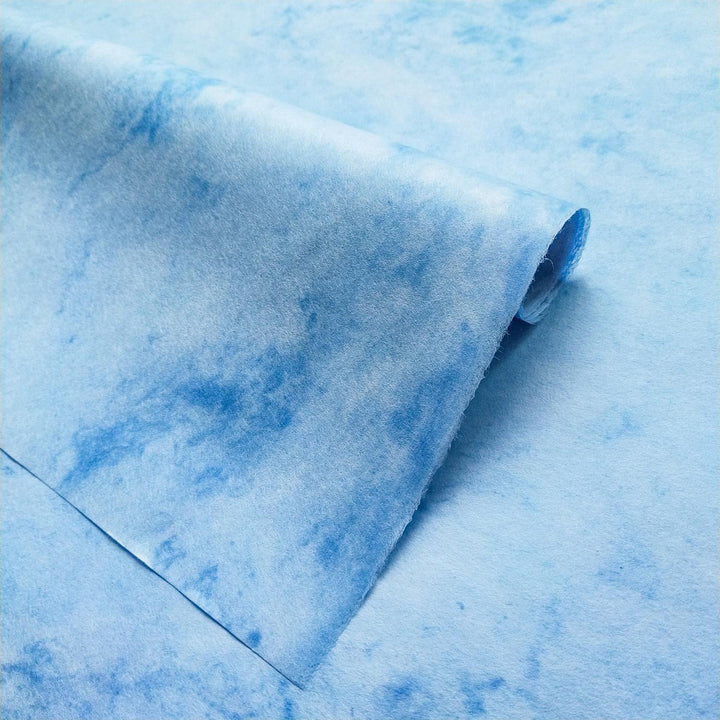 Marbled sky-blue mulberry paper by Kozo Studio with subtle white textures and a natural curled fold, ideal for artistic and craft applications.