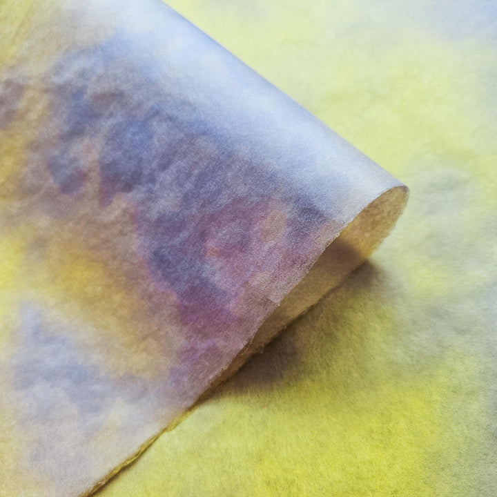 A close-up of the Ombre Kozo Mulberry Paper (#3 Canary Glow) by Kozo Studio showcases swirling yellow, purple, and gray hues. The semi-translucent texture and soft gradient create an artistic, abstract look with a slightly rolled edge.