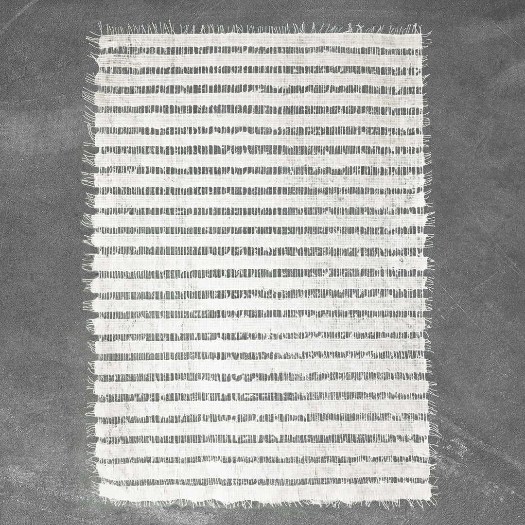 A small rectangular, rice paper-like rug with white and gray horizontal stripes from Kozo Studio. Its delicately fringed edges rest gracefully on a smooth, dark gray surface, reminiscent of the Handmade Geloma Kozo Paper (White).
