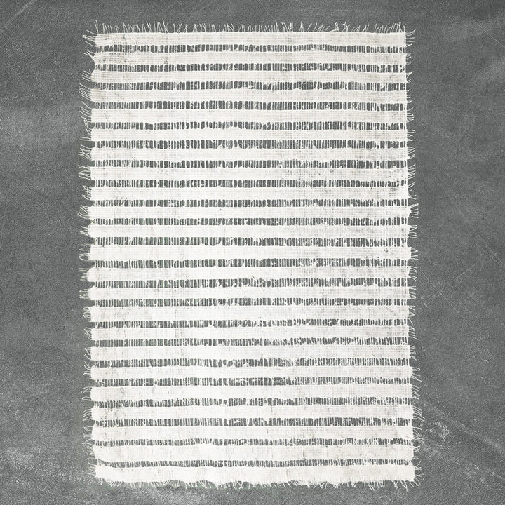 A small rectangular, rice paper-like rug with white and gray horizontal stripes from Kozo Studio. Its delicately fringed edges rest gracefully on a smooth, dark gray surface, reminiscent of the Handmade Geloma Kozo Paper (White).