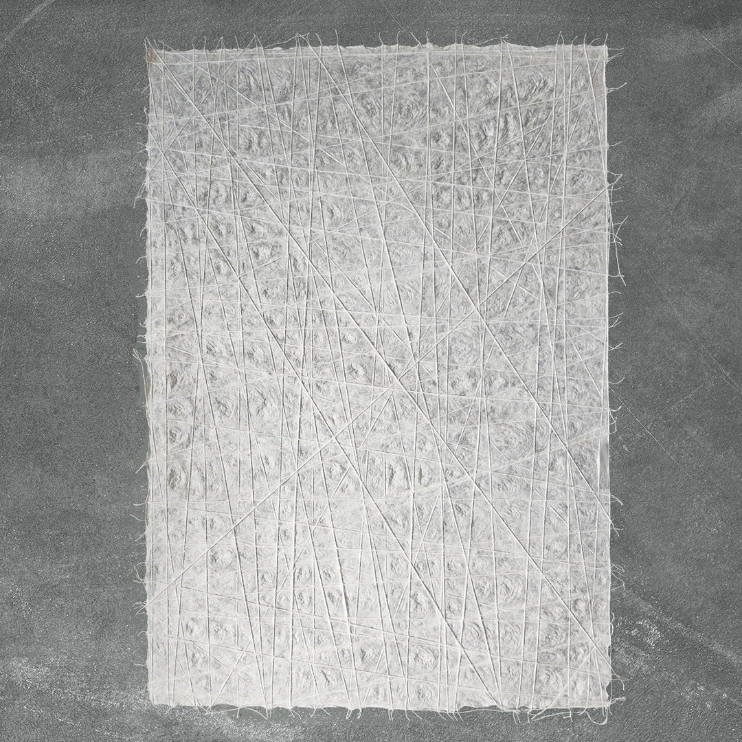 A rectangular Handmade Interlace Kozo Paper (White) from Kozo Studio, featuring an intricate pattern of thin, crisscrossing white lines and a rough, fibrous texture, is set against a plain dark gray background.