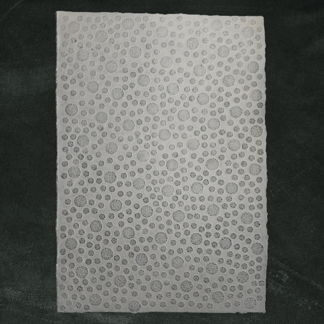 The Handmade Lace Kozo Paper (Bubble) from Kozo Studio, featuring slightly varying circular perforations, is set against a soft dark green fabric, offering a textured white rice paper with an intricate design.