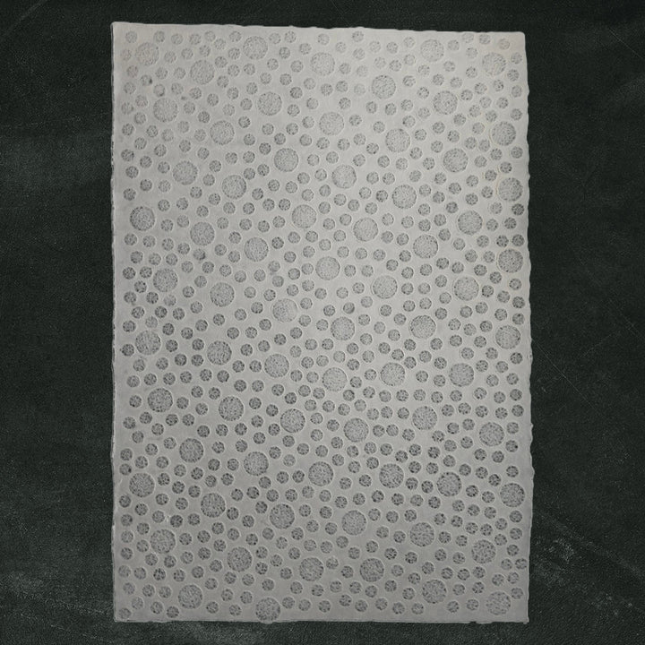 The Handmade Lace Kozo Paper (Bubble) from Kozo Studio, featuring slightly varying circular perforations, is set against a soft dark green fabric, offering a textured white rice paper with an intricate design.