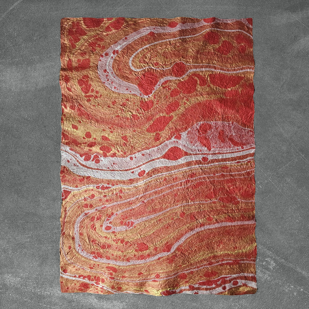 A sheet of Milano Kozo Mulberry Paper - Design 14 (Red) from Kozo Studio rests on a textured gray surface, showcasing swirling patterns in shades of red, orange, white, and gold for a vibrant design reminiscent of natural stone or lava.