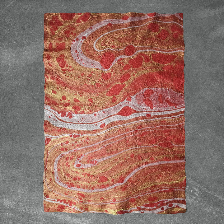 A sheet of Milano Kozo Mulberry Paper - Design 14 (Red) from Kozo Studio rests on a textured gray surface, showcasing swirling patterns in shades of red, orange, white, and gold for a vibrant design reminiscent of natural stone or lava.