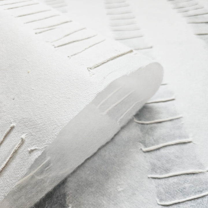 The Handmade Fluo Kozo Paper (White) by Kozo Studio offers a close-up view of textured mulberry sheets with fringed edges, showcasing a handcrafted look. Visible fibers and a soft, matte finish add depth and subtle shadows to the layered sheets.