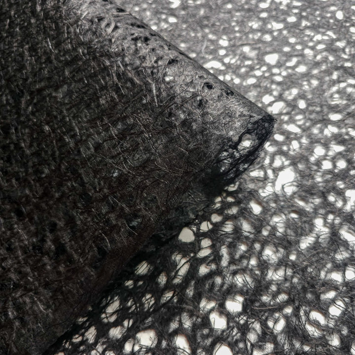 Close-up of Kozo Studios Asarakusui Lace Paper (Black) roll, offering a fibrous, semi-transparent texture of mulberry paper with an intricate lace-like design and irregular holes, spread on a surface.