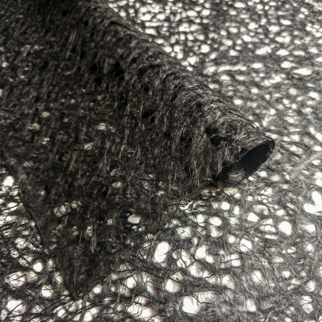 Close-up of Asarakusui Lace Paper (Black) by Kozo Studio, showcasing its textured netting of delicate mulberry paper, with intricate, irregular patterns and a lacy appearance. The partially unrolled lace paper reveals a sheer surface with various gaps and holes.