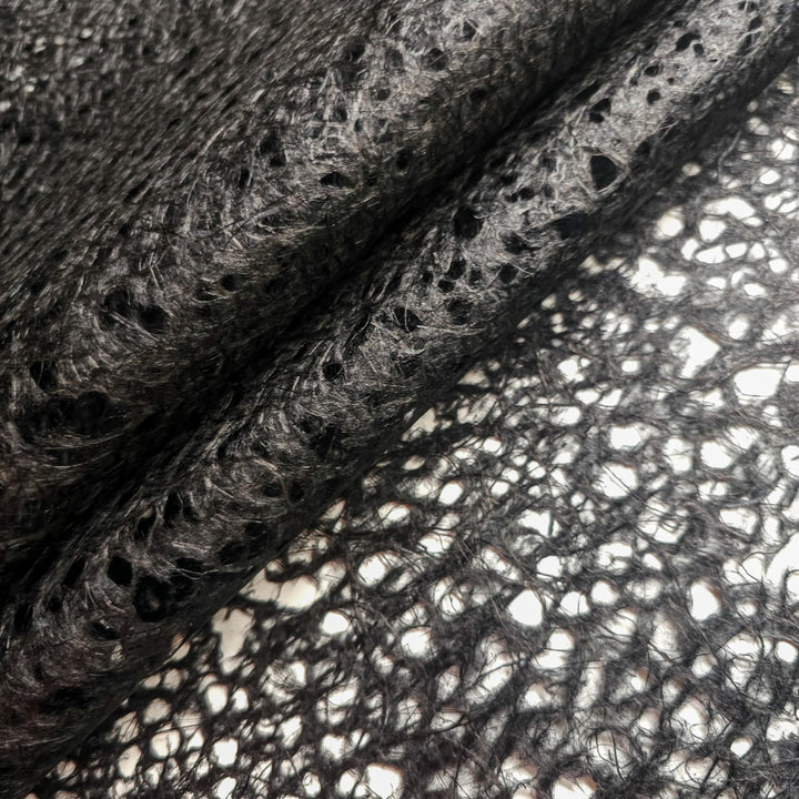 A close-up of the Asarakusui Lace Paper (Black) by Kozo Studio reveals intertwined black lace with rice paper fibers. The intricate patterns and holes, elegantly folded against a light background, highlight the harmonious blend of materials.