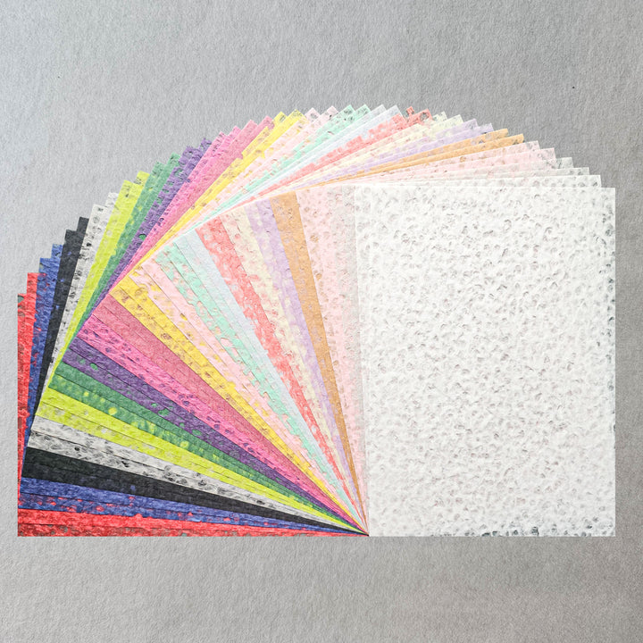 The Kozo Studio Assorted A4 Rakusui Mulberry Paper Set (40 Sheets) features colorful textured sheets fanned out on a gray surface, with a white sheet shown fully—ideal for decorative craft projects.