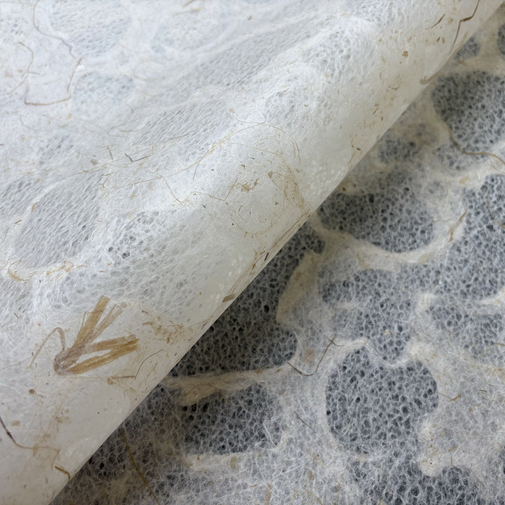 A close-up of Kozo Studio’s Tidal Lace Kozo Paper (Banana Fiber) shows its textured, translucent surface with organic patterns and embedded natural fibers, partially lifted to reveal the speckled layer beneath, showcasing its eco-friendly, handmade look.