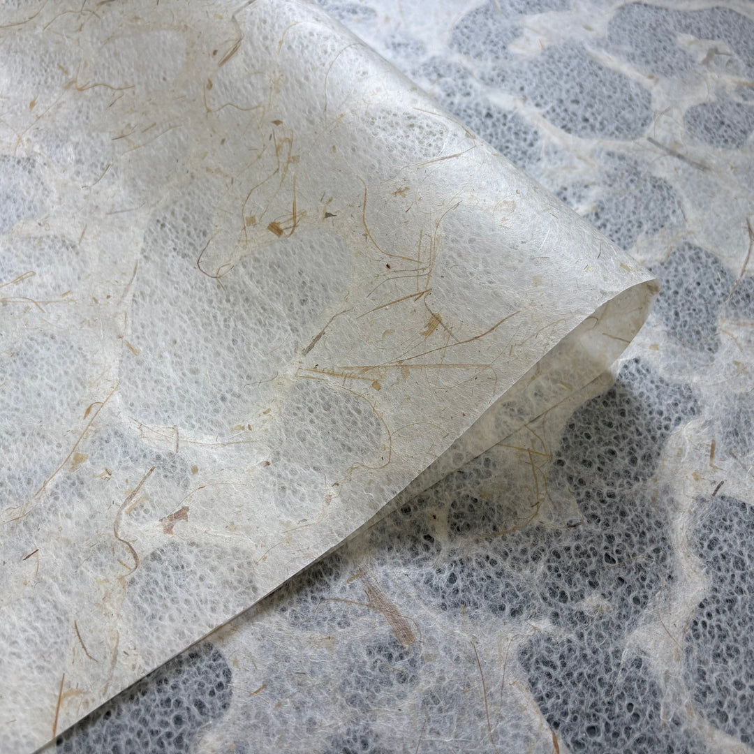A close-up of Kozo Studios Tidal Lace Kozo Paper (Banana Fiber) shows its textured surface, natural fibers, and translucent areas, partially folded atop another sheet of the same material.