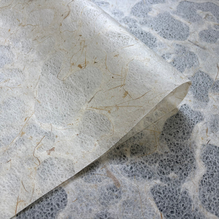 A close-up of Kozo Studios Tidal Lace Kozo Paper (Banana Fiber) shows its textured surface, natural fibers, and translucent areas, partially folded atop another sheet of the same material.