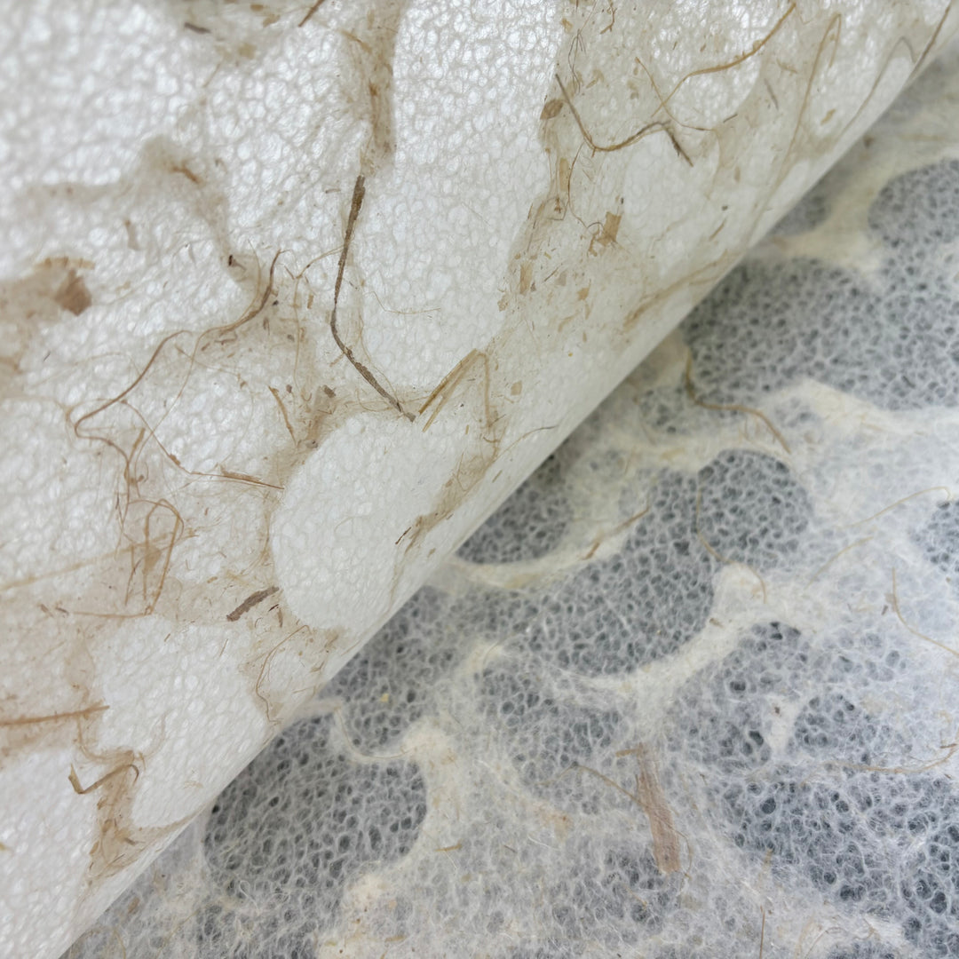 Close-up of Kozo Studios Tidal Lace Kozo Paper (Banana Fiber), showing delicate brown and white fibers and organic patterns. The partially rolled eco-friendly sheet reveals similar texture underneath, highlighting its natural, artisanal details.