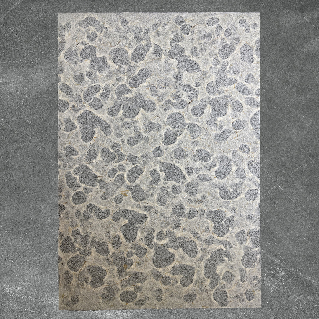 A rectangular sheet of Kozo Studios Tidal Lace Kozo Paper (Banana Fiber), featuring an irregular pebble-like gray pattern, is displayed on a slightly darker textured gray surface.