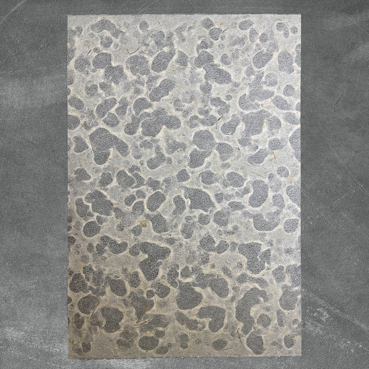 A rectangular sheet of Kozo Studios Tidal Lace Kozo Paper (Banana Fiber), featuring an irregular pebble-like gray pattern, is displayed on a slightly darker textured gray surface.