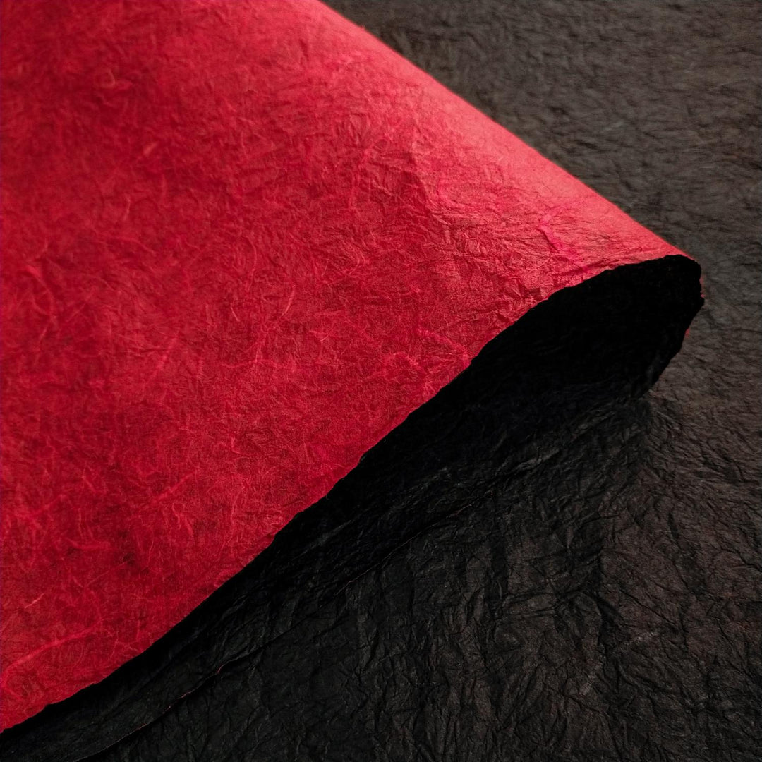 Close-up of Kozo Studio double-sided Momigami mulberry paper with a red sheet curled to reveal the black handmade paper beneath, highlighting its crinkled texture.