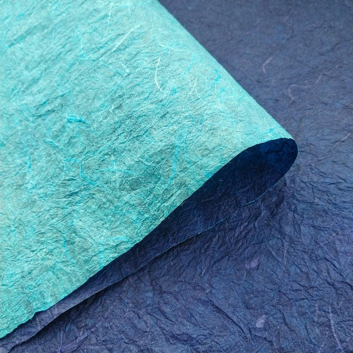 Close-up of handmade mulberry paper by Kozo Studio, featuring a curled light blue sheet of textured double-sided Momigami Kozo Paper on a dark blue background.