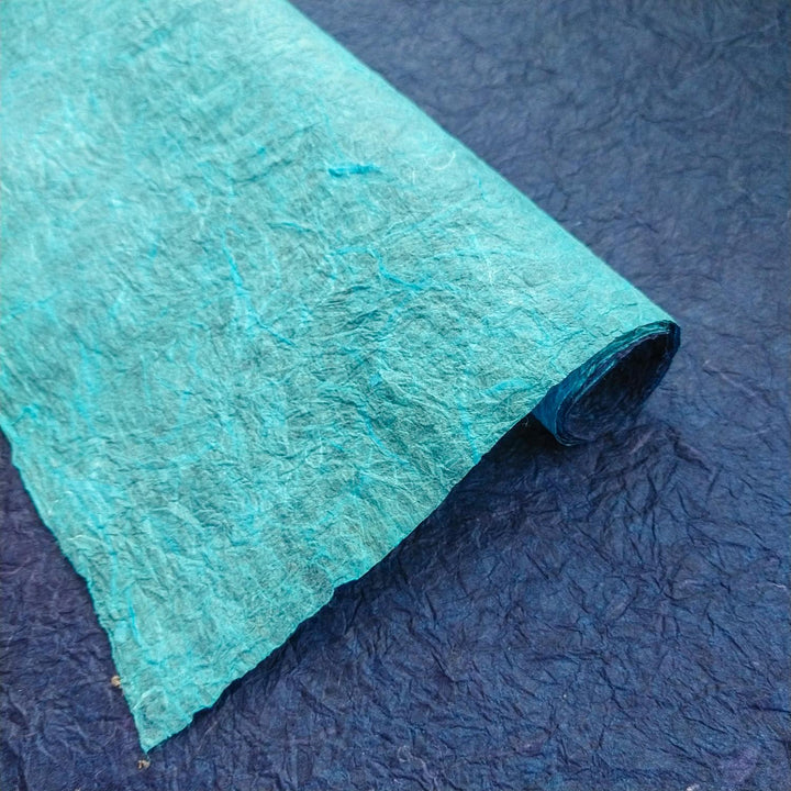 Artistic composition of double-sided Momigami Kozo Paper by Kozo Studio, showing layered mulberry paper with a light blue surface against a dark blue background.