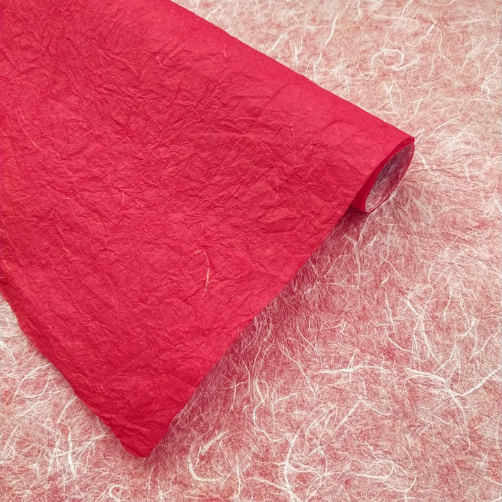 Kozo Studio's double-sided handmade mulberry paper, featuring red and white hues, rests rolled over a red surface, showcasing its rich fiber patterns and depth.