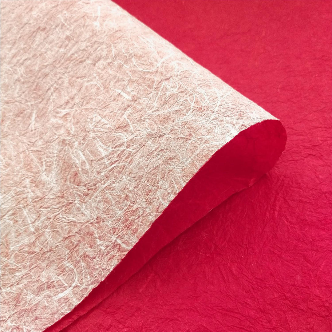 A double-sided sheet of handmade mulberry paper in white and red by Kozo Studio is gently rolled on a red background, highlighting its crinkled texture and fibrous detail.