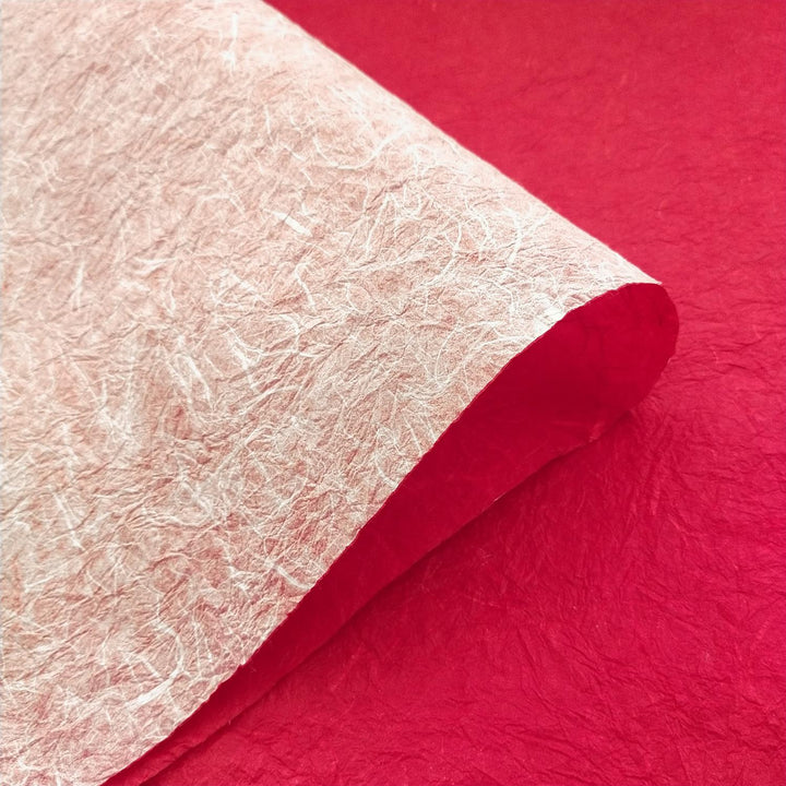 A double-sided sheet of handmade mulberry paper in white and red by Kozo Studio is gently rolled on a red background, highlighting its crinkled texture and fibrous detail.