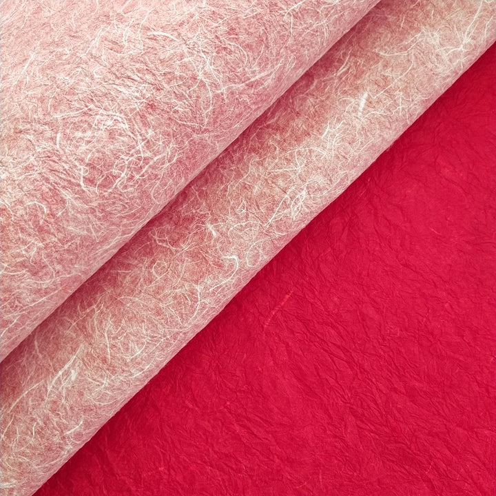 Rolled handmade mulberry paper in white and red by Kozo Studio sits on a solid red surface, showing off its textured layers and artistic appeal.