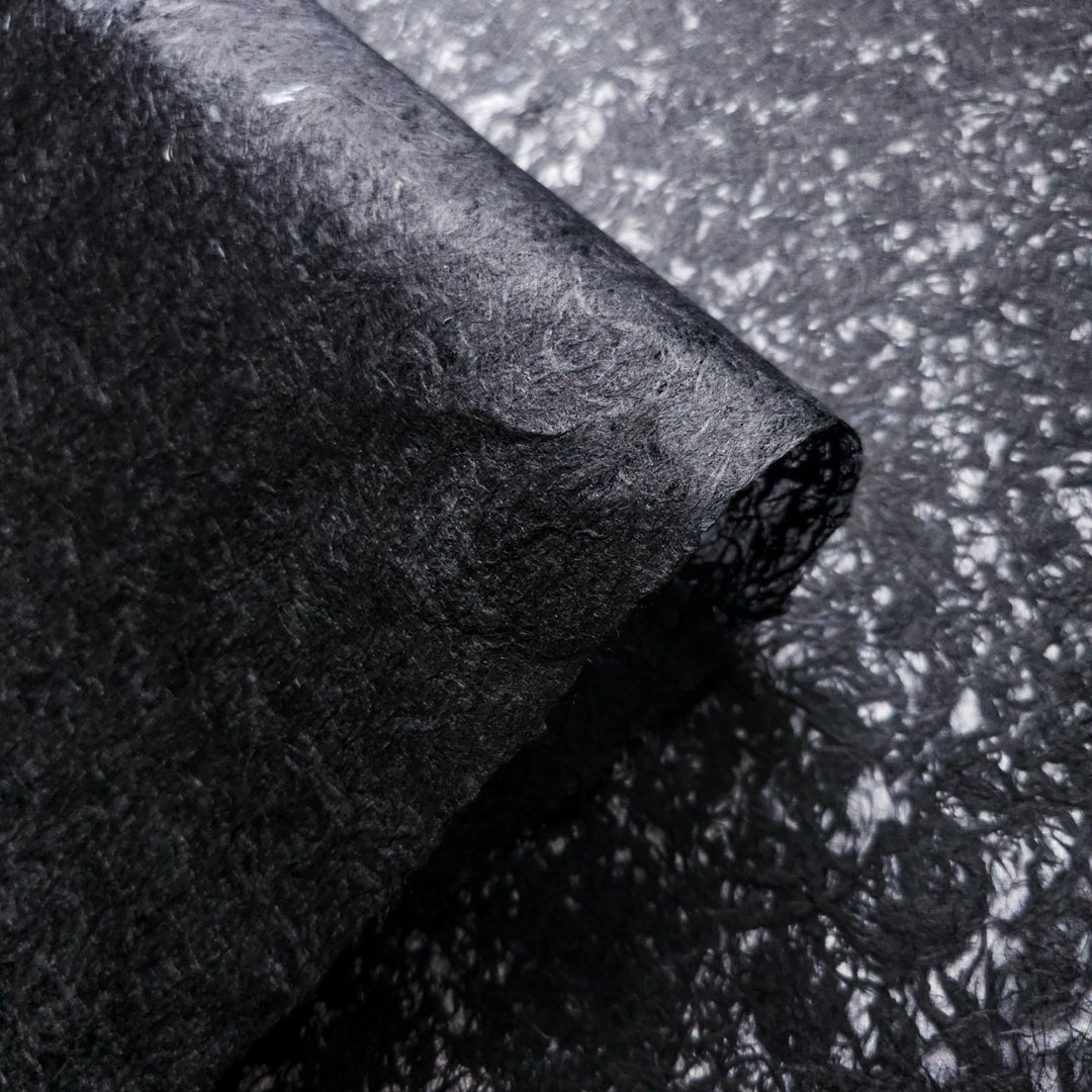 A close-up of Kozo Studios Fiber Veil Kozo Paper (Black) reveals a dark, textured, fibrous surface with intricate patterns on mulberry paper. The matte finish shows a section smoothly bending over itself, reminiscent of carbon fiber composites.