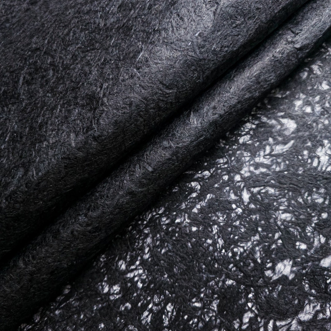 Close-up of Fiber Veil Kozo Paper (Black) by Kozo Studio, featuring a textured, fibrous mulberry surface. The top is soft and matte, while the bottom has a shinier finish with intricate patterns, creating a contrasting texture and sheen.