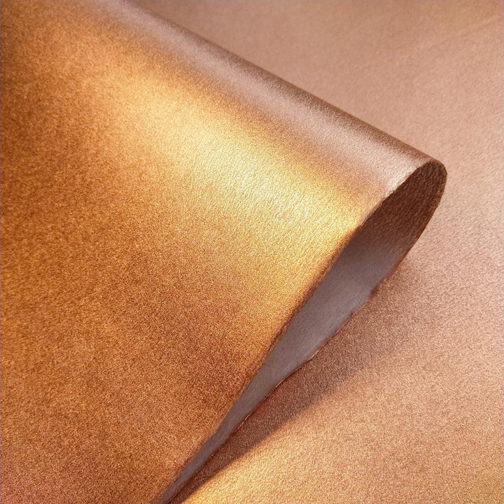 Partially unrolled sheet of shimmering copper handmade paper crafted by Kozo Studio, showing its unique texture and artisan quality.