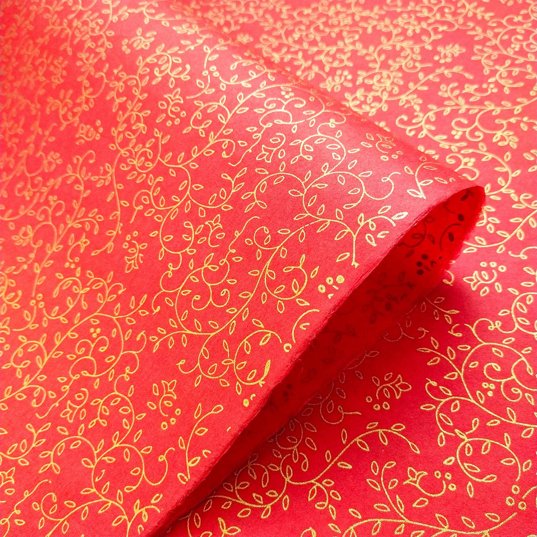 The Grapevine Screen-printed Kozo Paper Red by Kozo Studio is partially unrolled, revealing its intricate gold floral and vine patterns on fine mulberry paper.