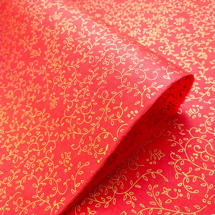 The Grapevine Screen-printed Kozo Paper Red by Kozo Studio is partially unrolled, revealing its intricate gold floral and vine patterns on fine mulberry paper.