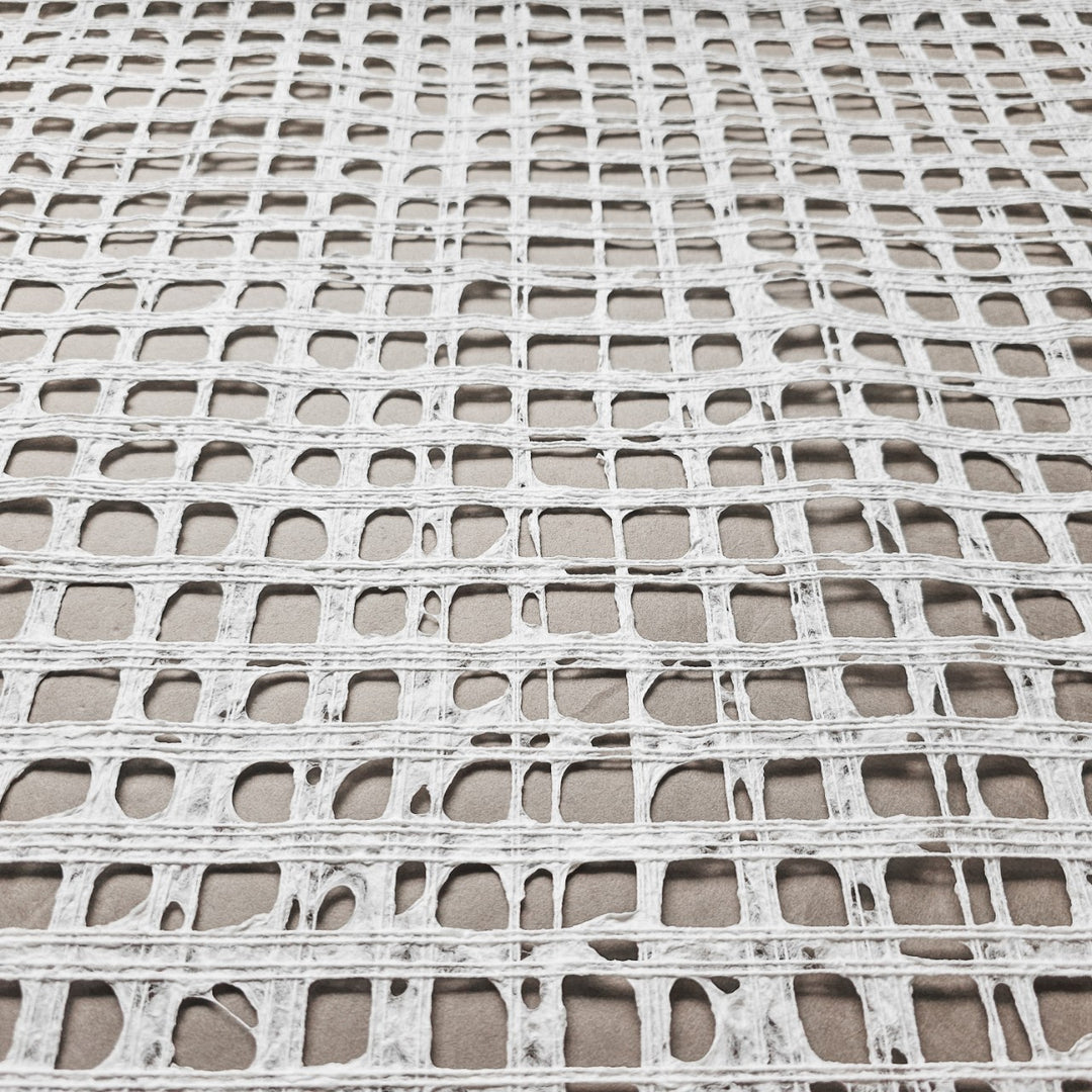 A close-up of the Handmade Barcelona Kozo Paper (White) by Kozo Studio shows a textured, grid-like pattern crafted from thin mulberry paper. The irregular rectangles and squares resemble a delicate net on a gray surface.