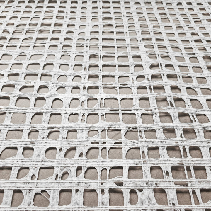 A close-up of the Handmade Barcelona Kozo Paper (White) by Kozo Studio shows a textured, grid-like pattern crafted from thin mulberry paper. The irregular rectangles and squares resemble a delicate net on a gray surface.